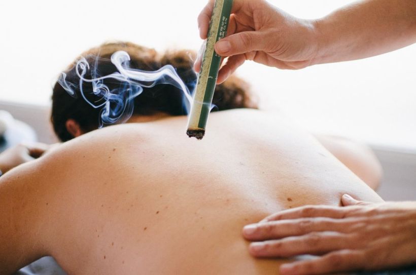 mujer de espaldas, torso desnudo, recibiendo una sesión de moxibustión. terapeuta sostiene en su mano derecha un cigarro de moxa, terapias naturales