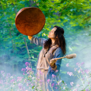 mujer rueda medicinal tocando tambor en el bosque rodeada de flores.